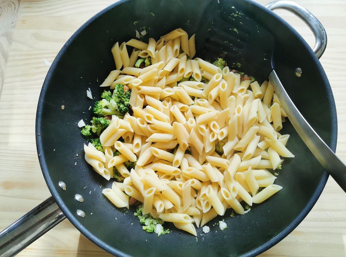 Cooked penne pasta with broccoli and pancetta in a frying pan.
