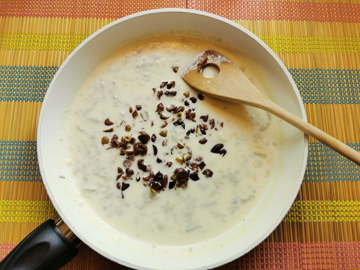 Chopped taggiasca olive added to creamy gorgonzola sauce in white pan.