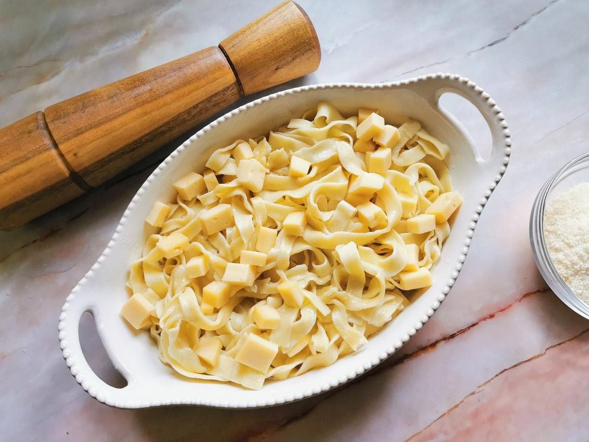 Cubes of fontina cheese on top of cooked tagliatelle in white oval oven bowl.