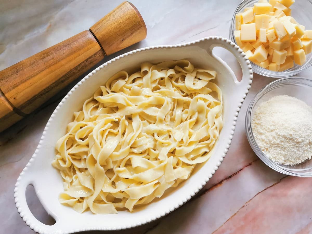 Cooked tagliatelle in white oval oven bowl and fontina cubes and grated Parmigiano in glass bowls.