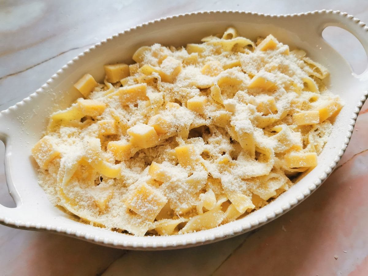 Cooked Tagliatelle and fontina cubes in white oval oven bowl with Parmigiano sprinkled on top.