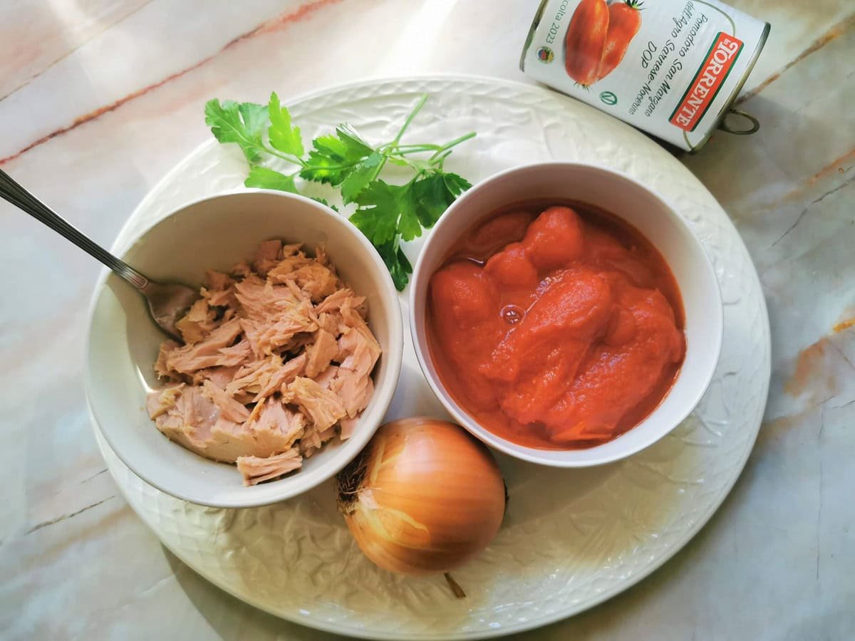 Ingredients for tuna bolognese sauce, can tuna in white bowl, peeled tomatoes in white bowl, twigs of parsley and an onion.